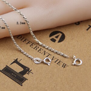 May include: Close-up of a silver-colored chain with a rope-like design. The chain is shown with a clasp and is approximately 1.7 mm to 2.3 mm in width. The background is a light brown color.