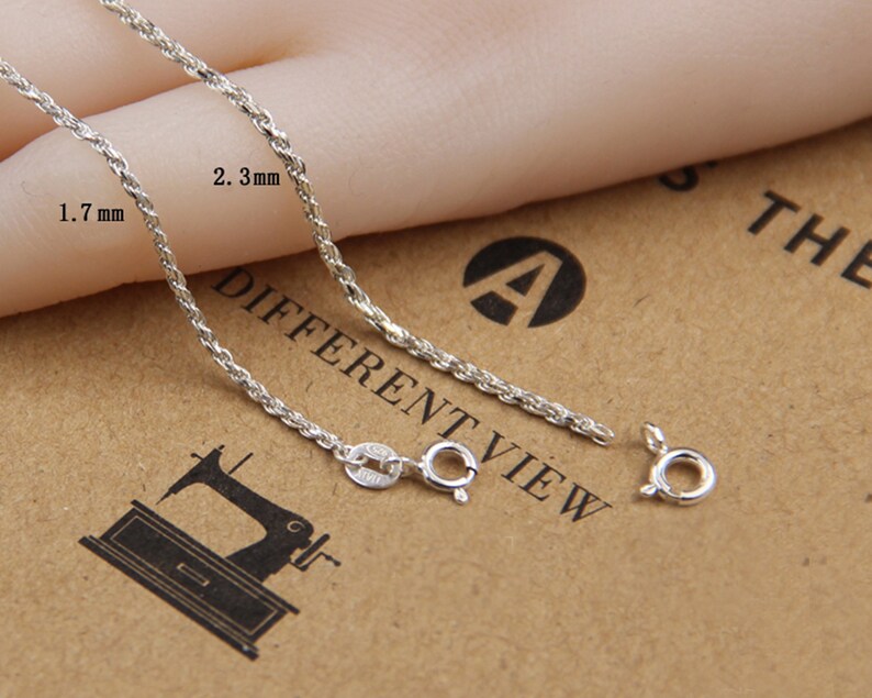 May include: Close-up of a silver-colored chain with a rope-like design. The chain is shown with a clasp and is approximately 1.7 mm to 2.3 mm in width. The background is a light brown color.