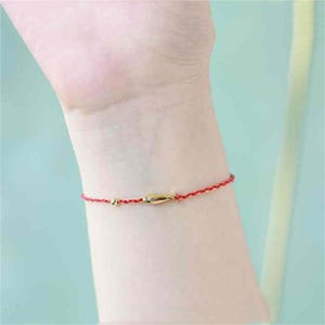 May include: A red string bracelet with a small gold fish charm. The bracelet is worn on a person's wrist.