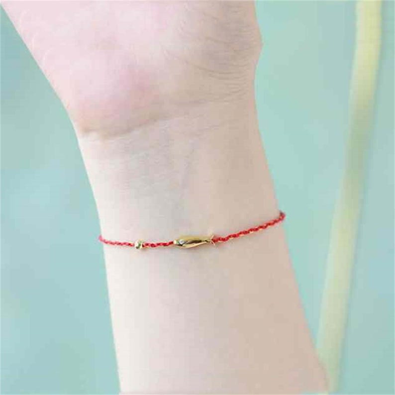 May include: A red string bracelet with a small gold fish charm. The bracelet is worn on a person's wrist.
