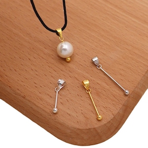May include: A collection of jewelry pendants on a wooden surface. The pendants include a large white pearl on a black cord, and several small pendants with a silver or gold finish, each with a small ball at the end.