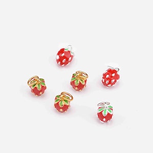 May include: A collection of small, red strawberry charms with white detailing and green leaves. Some have gold-coloured loops, others silver. The charms are scattered on a white background.