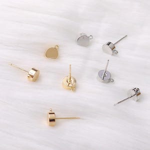 Può includere: Un assortimento di perni per orecchini color oro e argento con design piatti, rotondi e cilindrici. Ogni perno ha un piccolo anello per attaccare i componenti degli orecchini. Gli orecchini sono esposti su una superficie bianca e soffice.