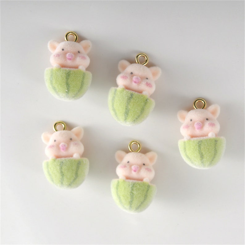 May include: Five pink pig charms with gold loops. Each pig is peeking out of a green watermelon-shaped charm.