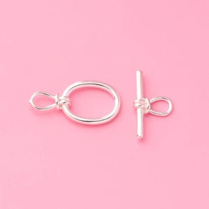 May include: A silver-toned toggle clasp for jewelry making, consisting of two separate components: a circular ring with an attached loop and a straight bar with a central loop, both featuring a smooth, polished metallic finish.