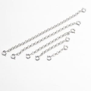 May include: Five silver chain extenders of different lengths, each with a spring ring clasp at one end and a small loop at the other. The chains are made of small, interconnected oval links. The background is plain white.
