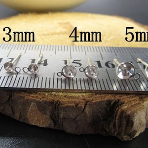 Può includere: Primo piano di diversi perni per orecchini color argento con piccole montature rotonde. I perni sono disposti in fila, con un righello che mostra le dimensioni di 3 mm, 4 mm e 5 mm. Gli orecchini sono su una superficie di legno.