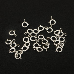 Puede incluir: Un grupo de anillos de salto de plata, también conocidos como anillos partidos, están dispersos sobre una superficie negra. Estos anillos se utilizan para conectar componentes de joyería.