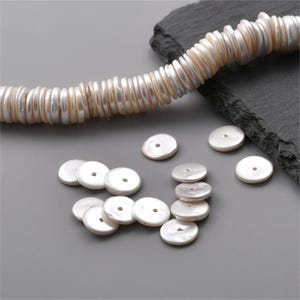 May include: A collection of flat, circular white mother-of-pearl beads with a central hole. Some beads are arranged in a long, curved strand, while others are scattered loosely. The beads have a smooth, iridescent surface and a lustrous finish.