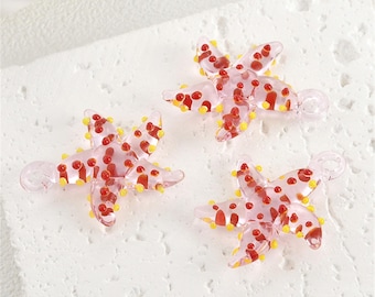 May include: Three clear glass starfish charms. Each charm is pink with red and yellow accents. The starfish have five points and a small loop for attaching to jewelry or other crafts. The charms are on a white surface.