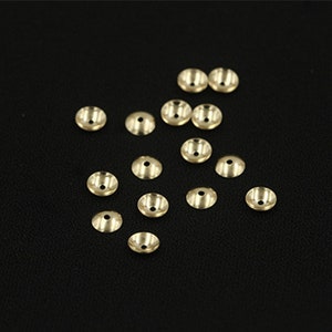 May include: A collection of 16 gold-colored metal bead caps, scattered on a black surface. The caps are round and have a slightly domed shape.