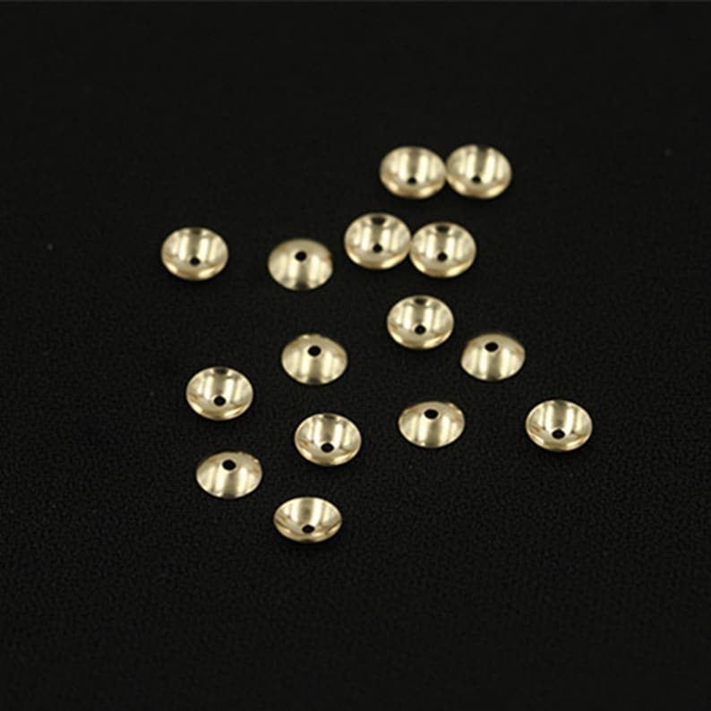 May include: A collection of 16 gold-colored metal bead caps, scattered on a black surface. The caps are round and have a slightly domed shape.