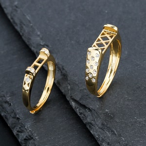Puede incluir: Dos anillos de oro con un diseño geométrico y piedras transparentes. Los anillos están sobre un fondo gris oscuro.