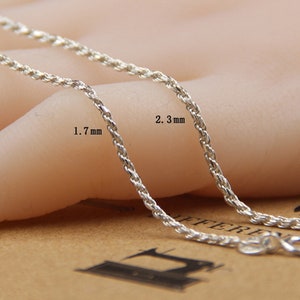 May include: Close-up of two silver-colored rope chains. One chain is 1.7 mm thick, and the other is 2.3 mm thick. The chains are displayed on a neutral background, showcasing their texture and design. The chains have a lobster clasp.