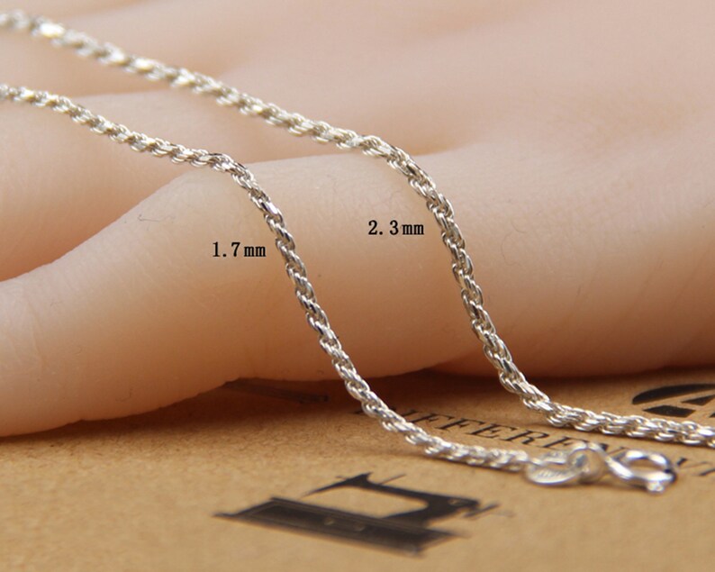 May include: Close-up of two silver-colored rope chains. One chain is 1.7 mm thick, and the other is 2.3 mm thick. The chains are displayed on a neutral background, showcasing their texture and design. The chains have a lobster clasp.