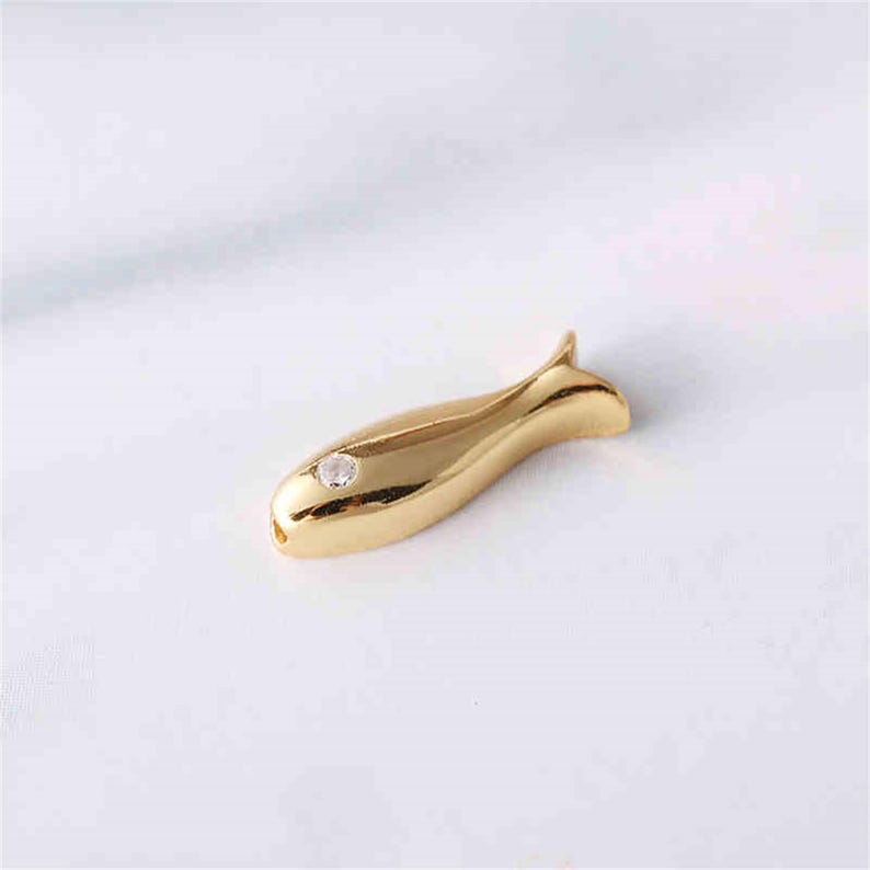 May include: A gold-coloured fish charm with a clear rhinestone in the centre. The charm is a popular choice for jewellery making and crafting.