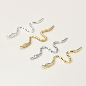 Op de afbeelding: Drie slangen-vormige bedels in zilver, goud en messing. Elke bedel heeft een kleine, heldere strasssteen op het hoofd.