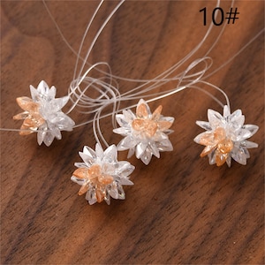 Puede incluir: Cinco abalorios de cristal transparente en forma de flor con un centro naranja claro. Los abalorios están unidos a finas cuerdas transparentes. Los abalorios están sobre una superficie de madera marrón. 10# está escrito en la esquina superior derecha de la imagen.