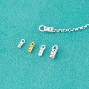 Puede incluir: Una colección de hallazgos de joyería plateados y dorados, que incluyen cierres y tapas finales, se muestra sobre un fondo turquesa. Los hallazgos están diseñados para la fabricación de joyas e incluyen una cadena.