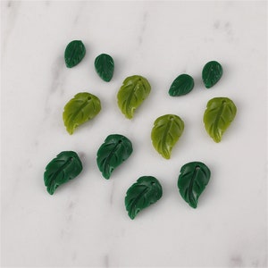 Könnte beinhalten: Eine Sammlung von 12 grünen, blattartigen Charms. Die Charms sind aus einem glatten, glänzenden Material gefertigt und haben ein realistisches Blattdesign.
