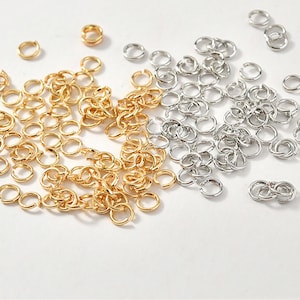 May include: A large quantity of small, gold and silver jump rings. The jump rings are scattered on a white surface.