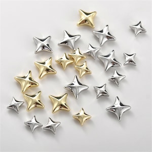 Puede incluir: Una colección de adornos en forma de estrella en tonos dorados y plateados. Las estrellas metálicas de varios tamaños están dispersas sobre una superficie blanca. Estos elementos decorativos podrían usarse para manualidades o joyería.
