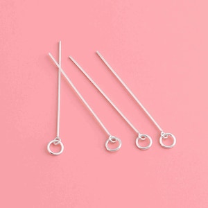 Puede incluir: Cuatro alfileres de cabeza plateados con un pequeño bucle en el extremo. Los alfileres están dispuestos en una fila sobre un fondo rosa.