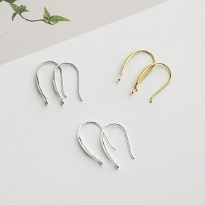 Puede incluir: Una colección de tres pares de ganchos para pendientes en tonos plateados y dorados. Los ganchos tienen un diseño curvo y minimalista. Los pendientes se muestran sobre una superficie blanca, con una ramita de vegetación en la esquina superior izquierda.