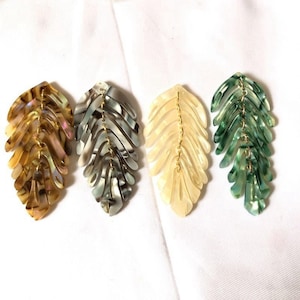 Op de afbeelding: Vier blad-vormige oorbellen in goud, grijs, crème en groen. Elke oorbel heeft een gelaagd ontwerp met een goudkleurige ketting in het midden. De oorbellen worden gepresenteerd op een wit oppervlak.