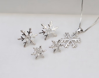 May include: A silver snowflake necklace and earrings set with clear crystal accents. The necklace features a larger snowflake pendant with two smaller snowflakes attached to it. The earrings are simple and feature a single snowflake each.
