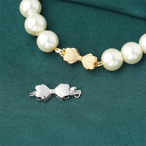 May include: A pearl necklace with a gold bow clasp and a silver bow clasp. The necklace is on a green fabric background.