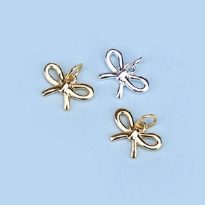 Op de afbeelding: Drie zilveren en gouden strikcharms. De charms zijn in de vorm van een strik met een lus aan de bovenkant om aan een ketting of armband te bevestigen.
