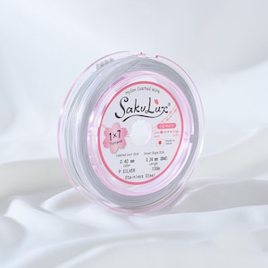May include: A spool of silver nylon-coated wire, branded as SakuLux, is displayed on a white surface. The label indicates a diameter of 0.40 mm and a length of 100m. The packaging is clear with pink accents and Japanese text.