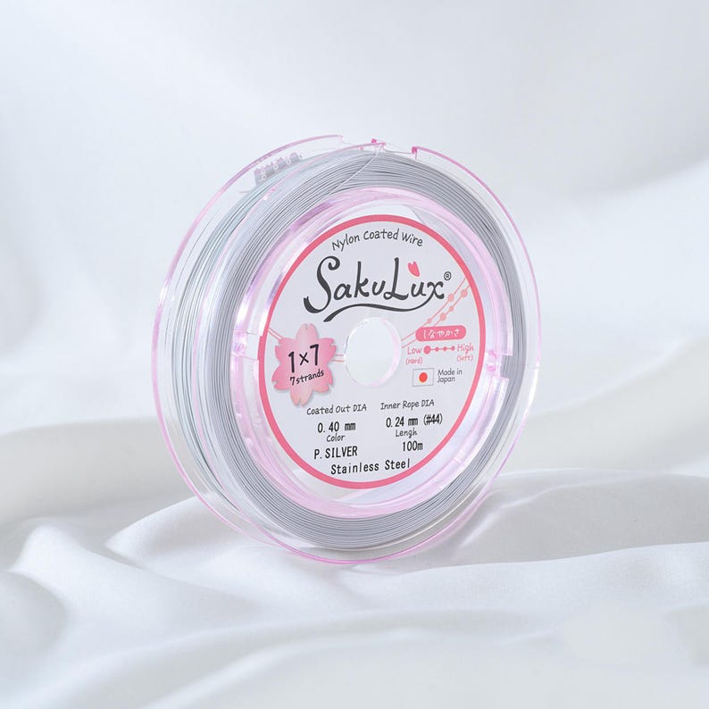 May include: A spool of silver nylon-coated wire, branded as SakuLux, is displayed on a white surface. The label indicates a diameter of 0.40 mm and a length of 100m. The packaging is clear with pink accents and Japanese text.