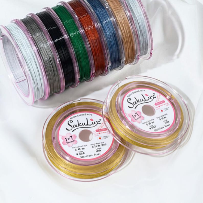 May include: Spools of nylon-coated wire in various colors, including gold, black, and silver. Two spools of gold wire are in clear plastic containers. The text "SakuLux" and "1x7" are visible on the labels.