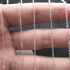 Può includere: Primo piano di diverse delicate collane a catena d'argento. Le catene sono sottili e presentano piccoli anelli interconnessi. Le catene sono esposte su uno sfondo neutro, evidenziando la loro superficie metallica riflettente.