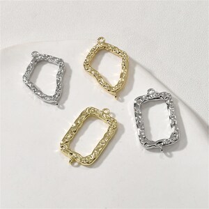 May include: Four metal frame charms, two gold and two silver, with a textured surface. The frames are rectangular with rounded corners and have a loop at the top for attaching to jewelry.