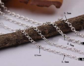 May include: Four silver chains of different thicknesses laid out on a brown wooden surface. The chains are labeled with their thickness in millimeters: 0.4 mm, 0.5 mm, 0.7 mm, 1.8 mm, and 2.0 mm.