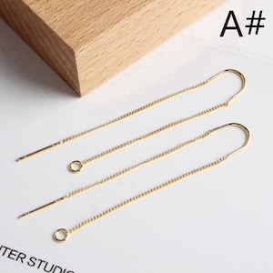 Könnte beinhalten: Goldfarbene Ohrringe zum Einfädeln. Die Ohrringe haben ein filigranes Kettendesign mit einem kleinen kreisförmigen Akzent. Die Oberseite des Ohrrings hat eine gebogene, hakenartige Form. Der Text "A#" ist ebenfalls zu sehen.