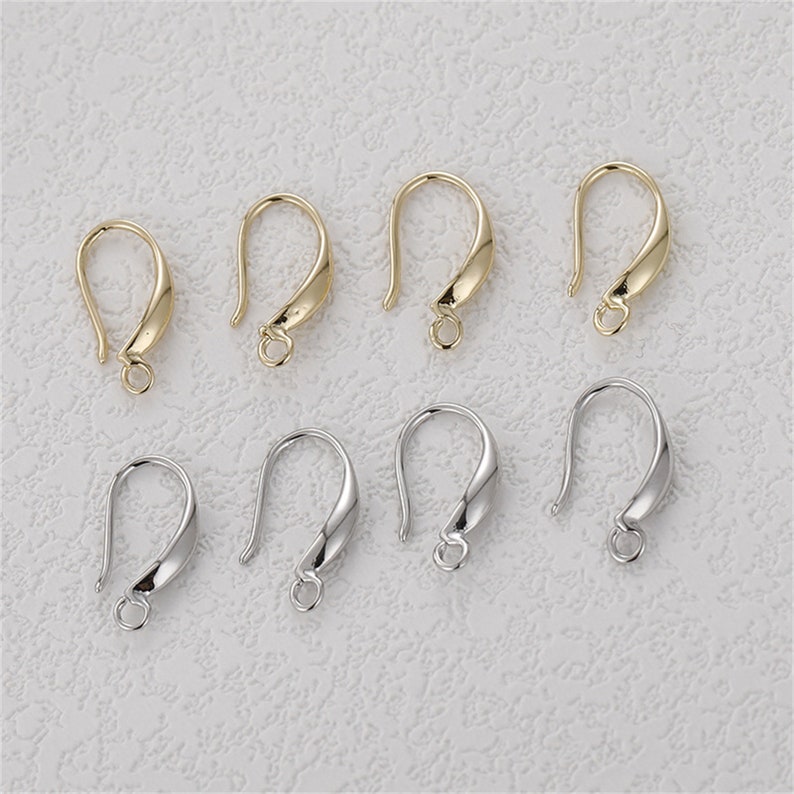May include: An assortment of gold and silver-colored earring hooks. The hooks are curved with a loop at the bottom for attaching charms or beads. The hooks are displayed on a textured, off-white surface.
