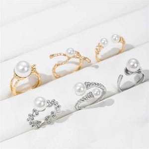 May include: Six rings with faux pearls in gold and silver tones. The rings have various designs, including a double pearl ring, a floral design, and a twisted band. The pearls are white and round.