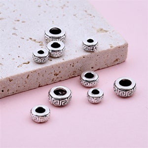 May include: A set of seven silver-toned metal beads with a geometric design. The beads are of varying sizes and are arranged on a light pink surface.