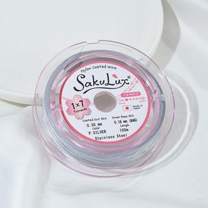 May include: A spool of SakuLux nylon-coated wire, 0.30 mm diameter, silver color, and 100m length. The package includes the text "1x7 7 strands" and "Stainless Steel". The wire is in a clear plastic container with a pink rim.