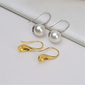 Peut inclure: Une paire de boucles d'oreilles avec une perle et un crochet en or. La perle est blanche et le crochet est un design simple et incurvé.