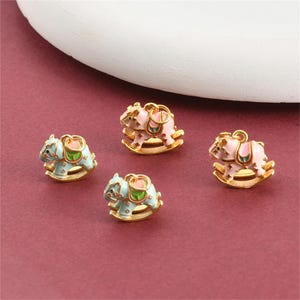 May include: Four gold-coloured rocking horse charms. Two are light blue with green and pink accents, and two are pink with green and pink accents. The charms are on a maroon surface.