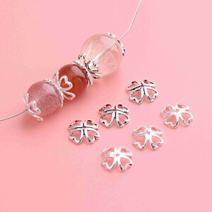 May include: Silver-toned metal bead caps in the shape of a four-leaf clover. The caps are scattered on a pink surface.