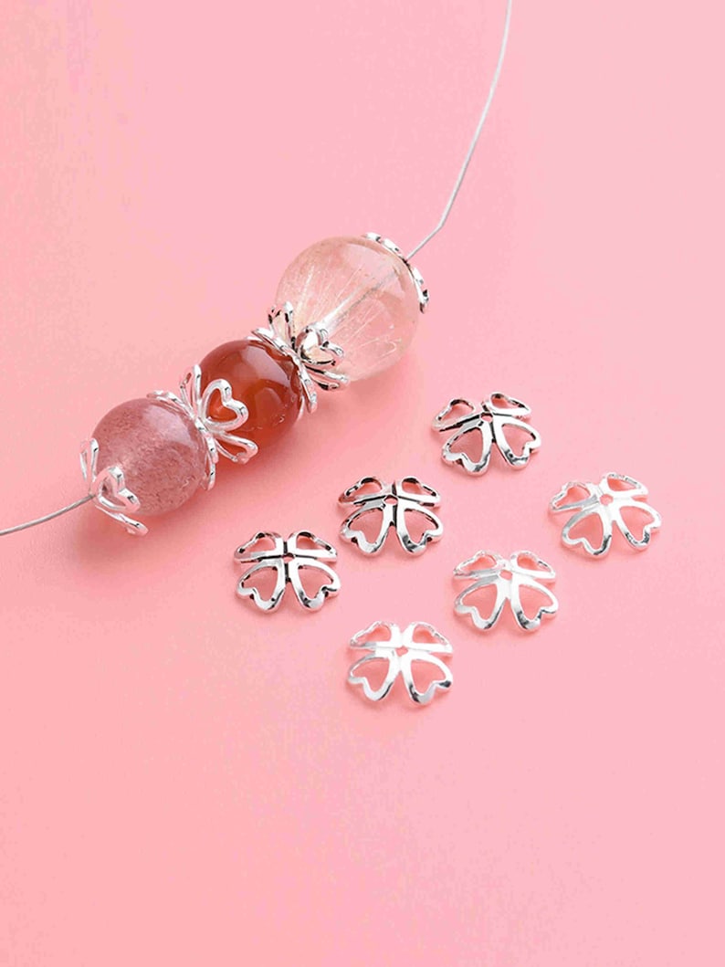 May include: Silver-toned metal bead caps in the shape of a four-leaf clover. The caps are scattered on a pink surface.