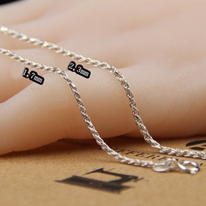May include: Two silver-colored chain necklaces, one 1.7 mm and the other 2.3 mm thick, are displayed. The chains have a twisted rope design and are shown with their clasps. The chains are on a light-colored background.