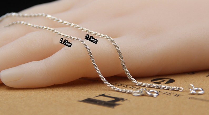 May include: Two silver-colored chain necklaces, one 1.7 mm and the other 2.3 mm thick, are displayed. The chains have a twisted rope design and are shown with their clasps. The chains are on a light-colored background.