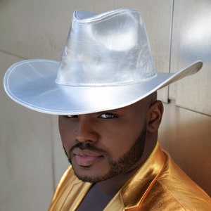 May include: A man wearing a silver cowboy hat and a gold metallic blazer.